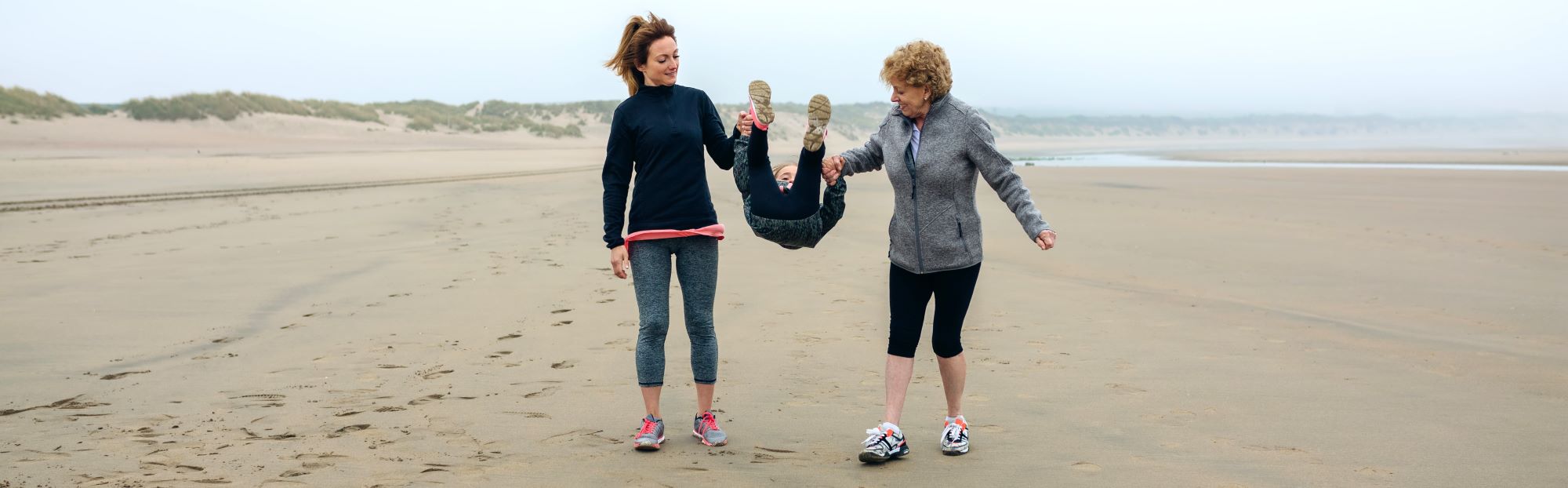 Three generations of females walking along a beach. Two older women are swinging the youngest in between them