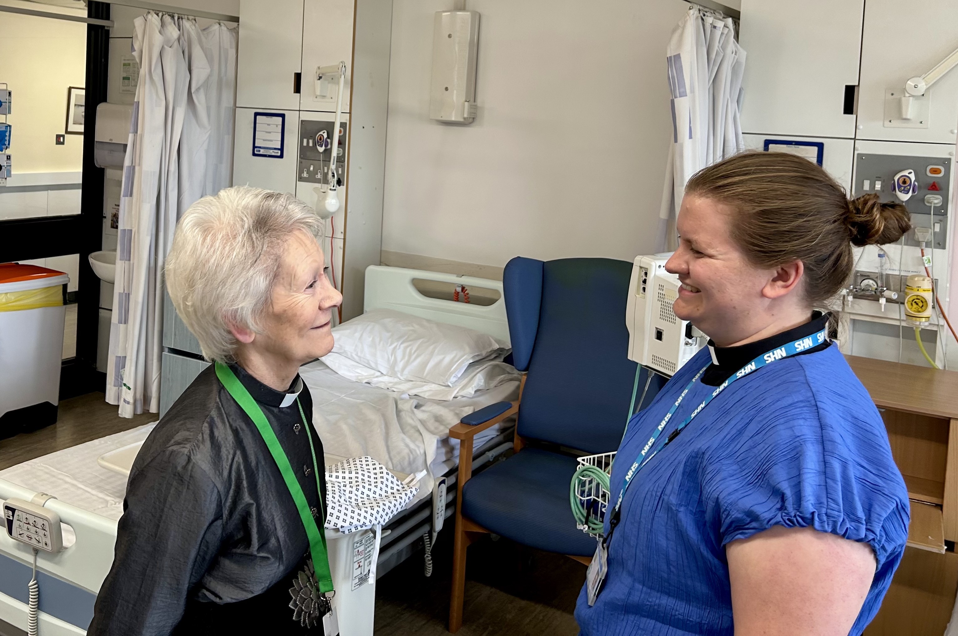 Reverend Christine Brown with Reverend Amy Bland standing in front of a hospital bed and looking towards eachother
