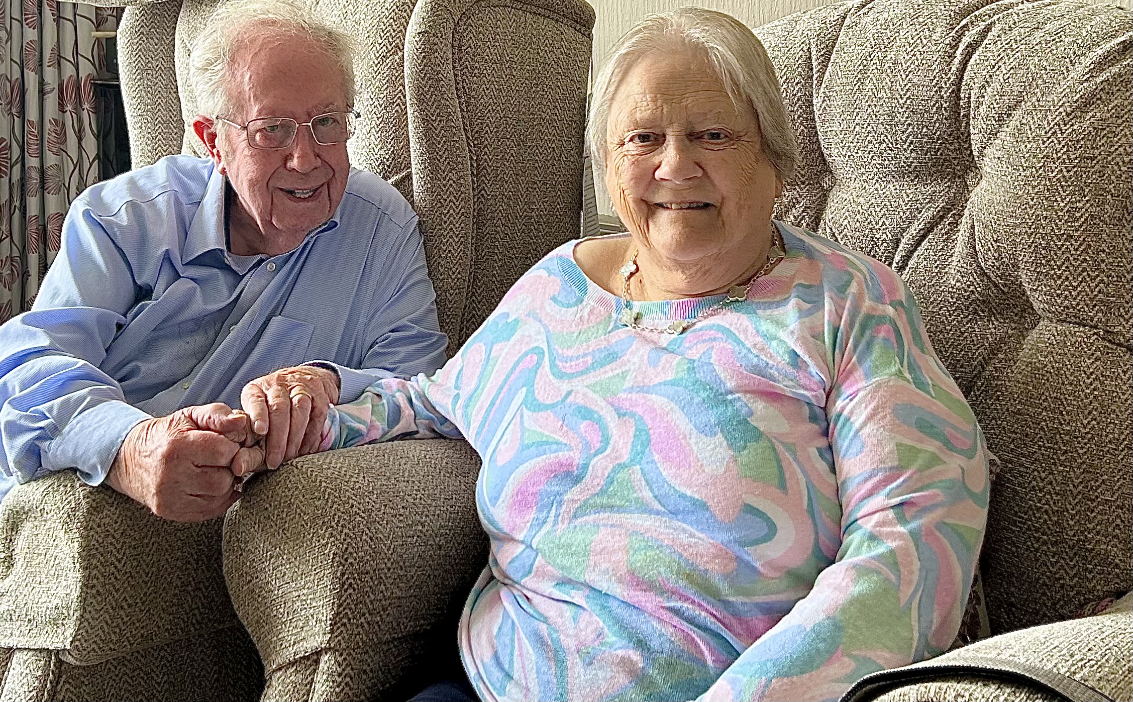 Eileen and John Unsworth sitting on a comfy sofa and holding hands while smiling to the camera